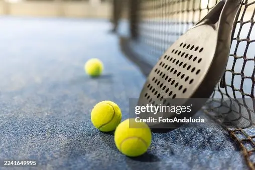 Padel racket resting beside three bright yellow tennis balls on a blue court.