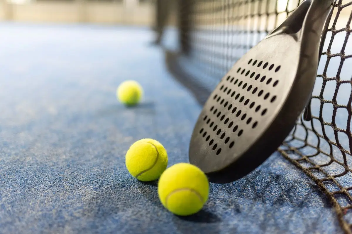 Padel racket resting beside three bright yellow tennis balls on a blue court.